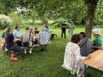Menschen sitzen auf Bierbänken an Tischen auf einer Wiese mit Bäumen