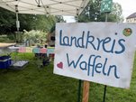 Ein Schild mit der Aufschrift "Landkreis Waffeln" vor einem Waffelstand auf einer Wiese 